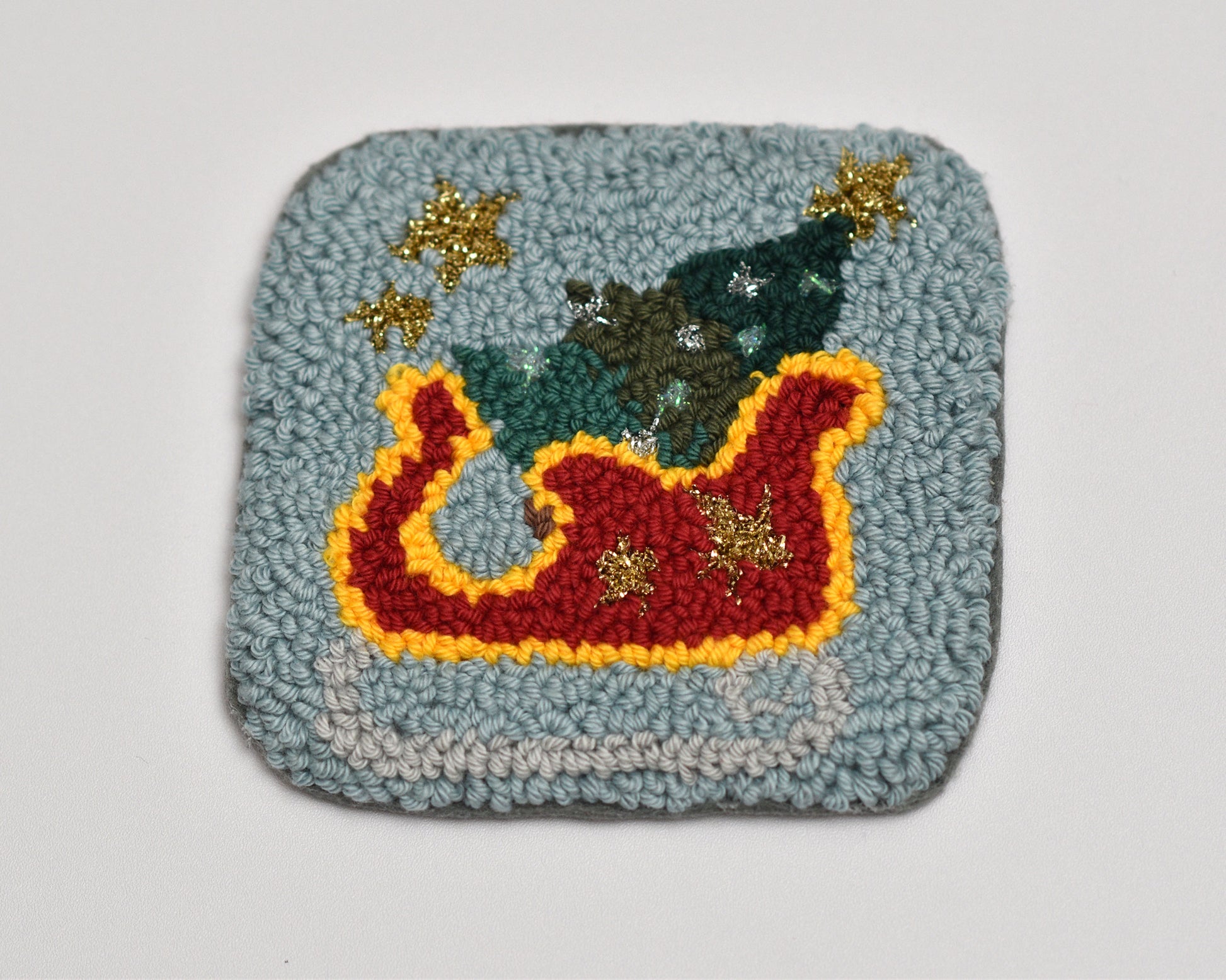 Textured square punch needle coaster with a colorful design of a sleigh, trees, and stars on a light gray background.