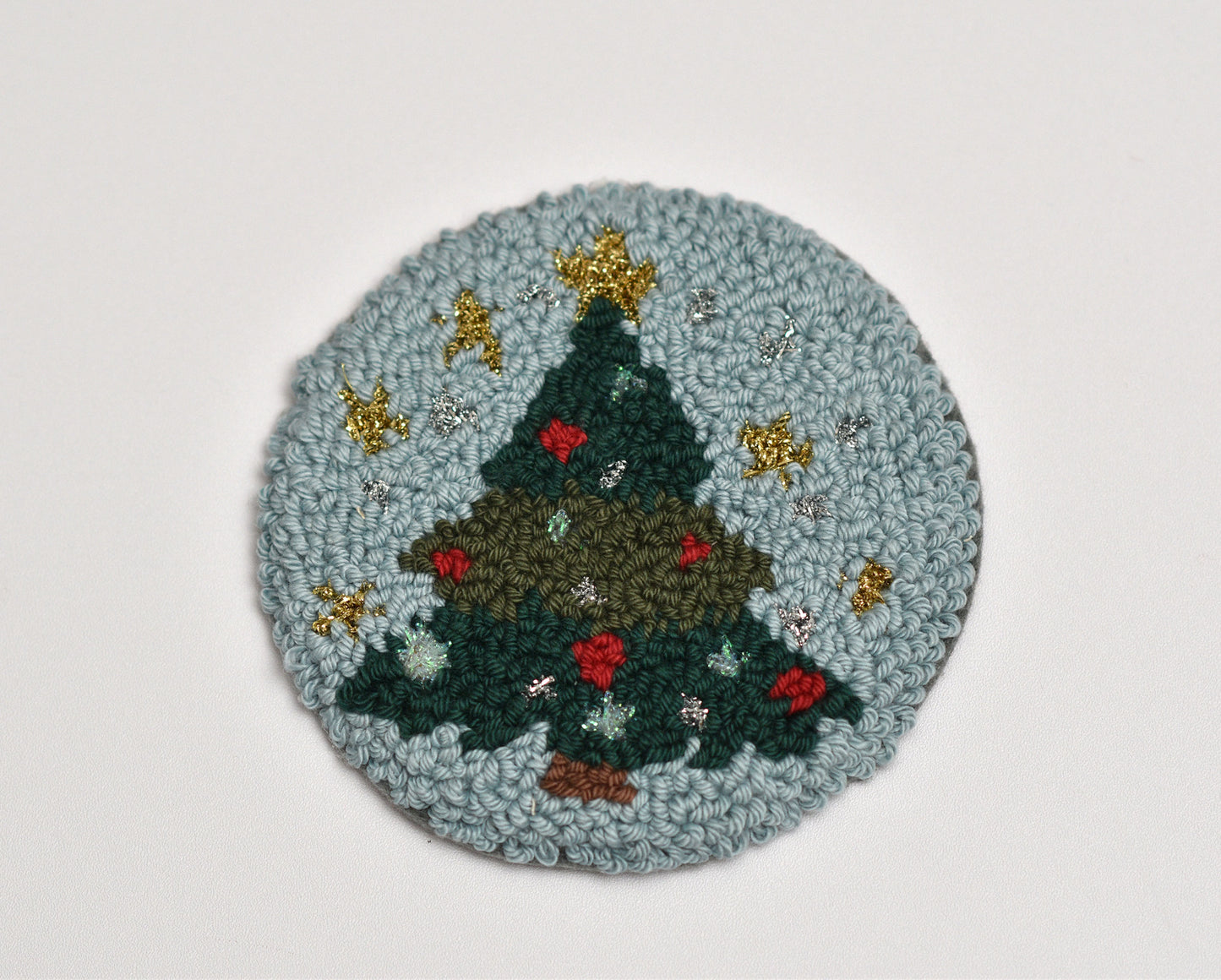 Round punch needle coaster with a Christmas tree design on a light gray background