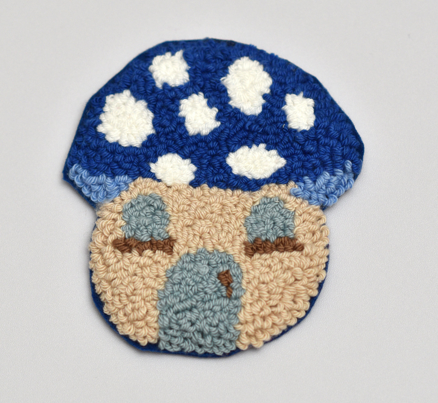 Punch needled mushroom coaster with blue cap and beige body on a light gray background
