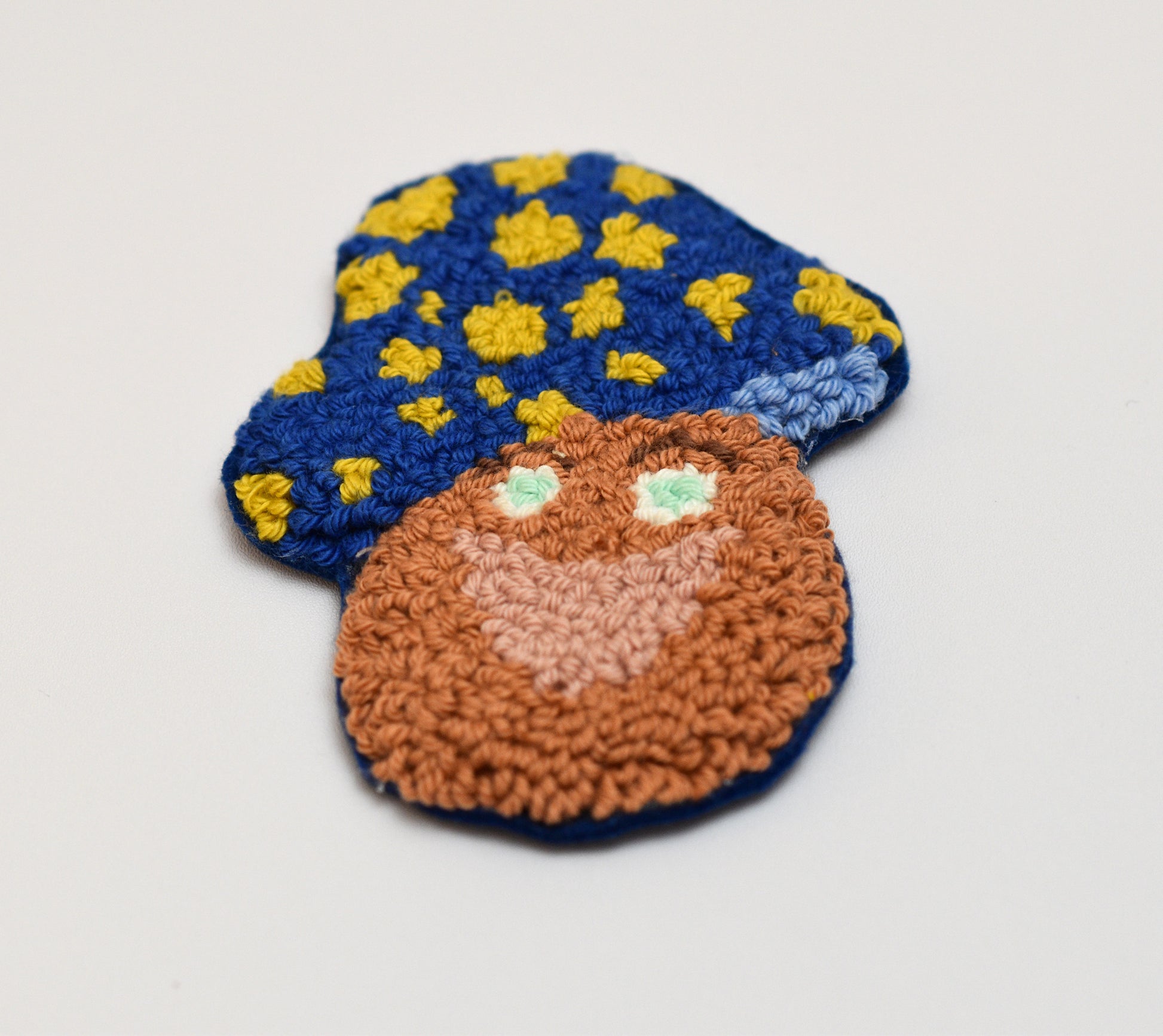 Punch needled mushroom shaped coaster with blue hat and yellow dots on a light gray background