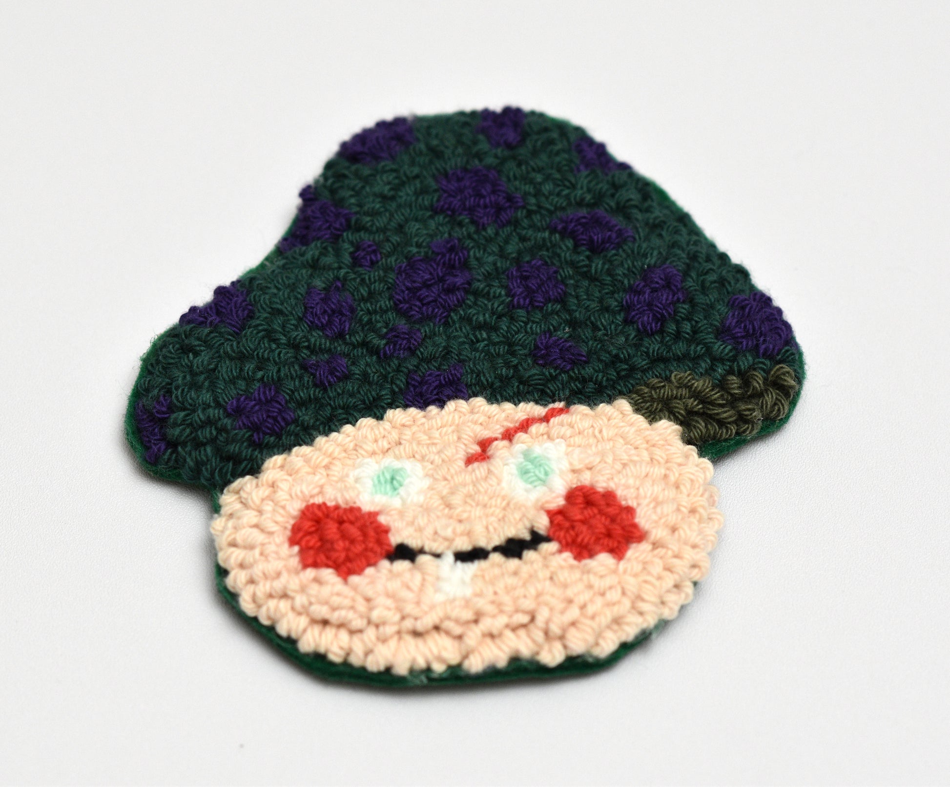 Punch needled coaster with mushroom character design with a green hat and beige face on a white background