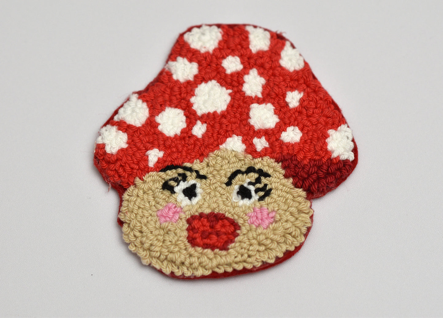 Punch needled coaster with design of a mushroom with a face on a light gray background