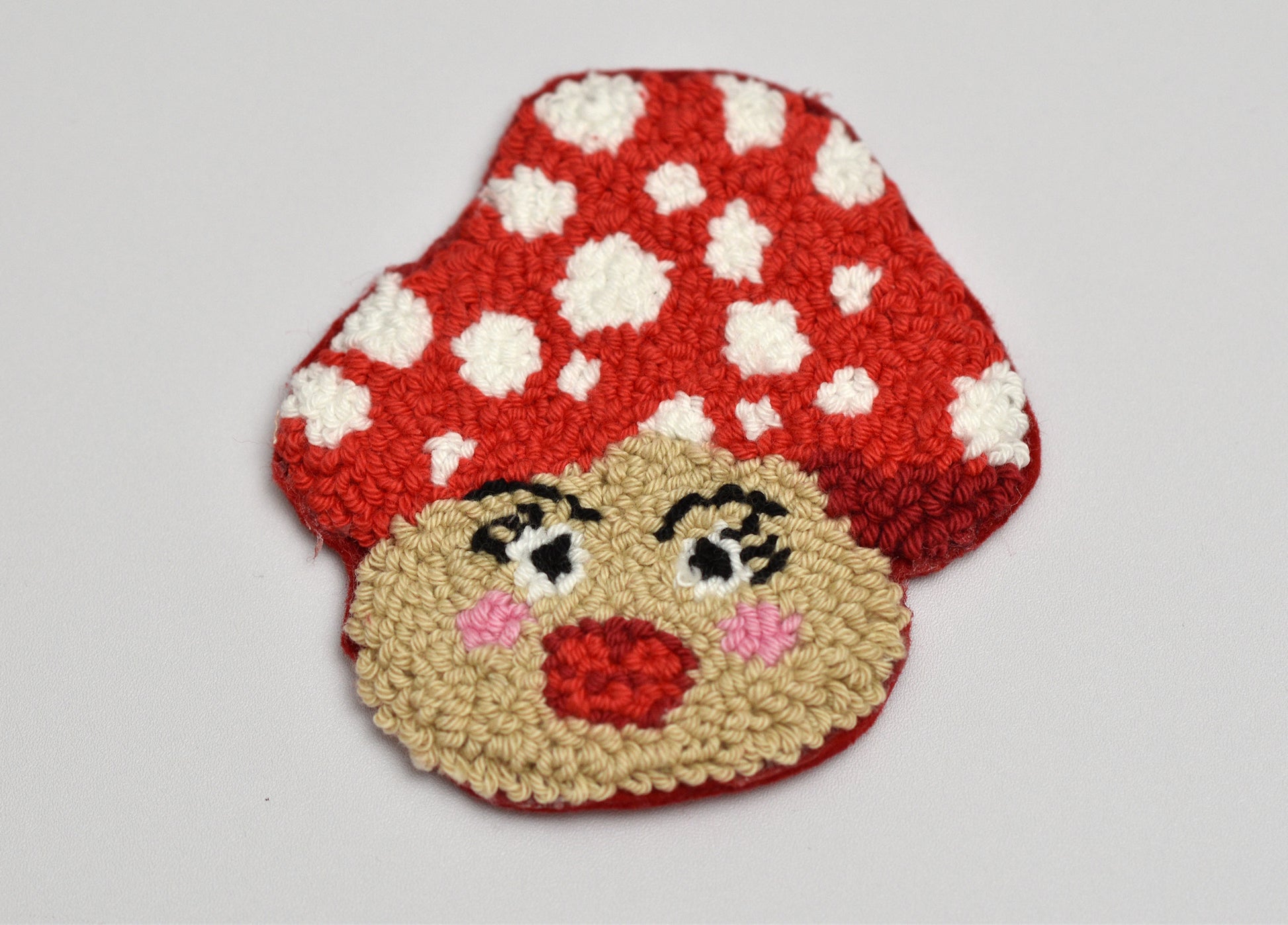 Punch needled coaster with design of a mushroom with a face on a light gray background