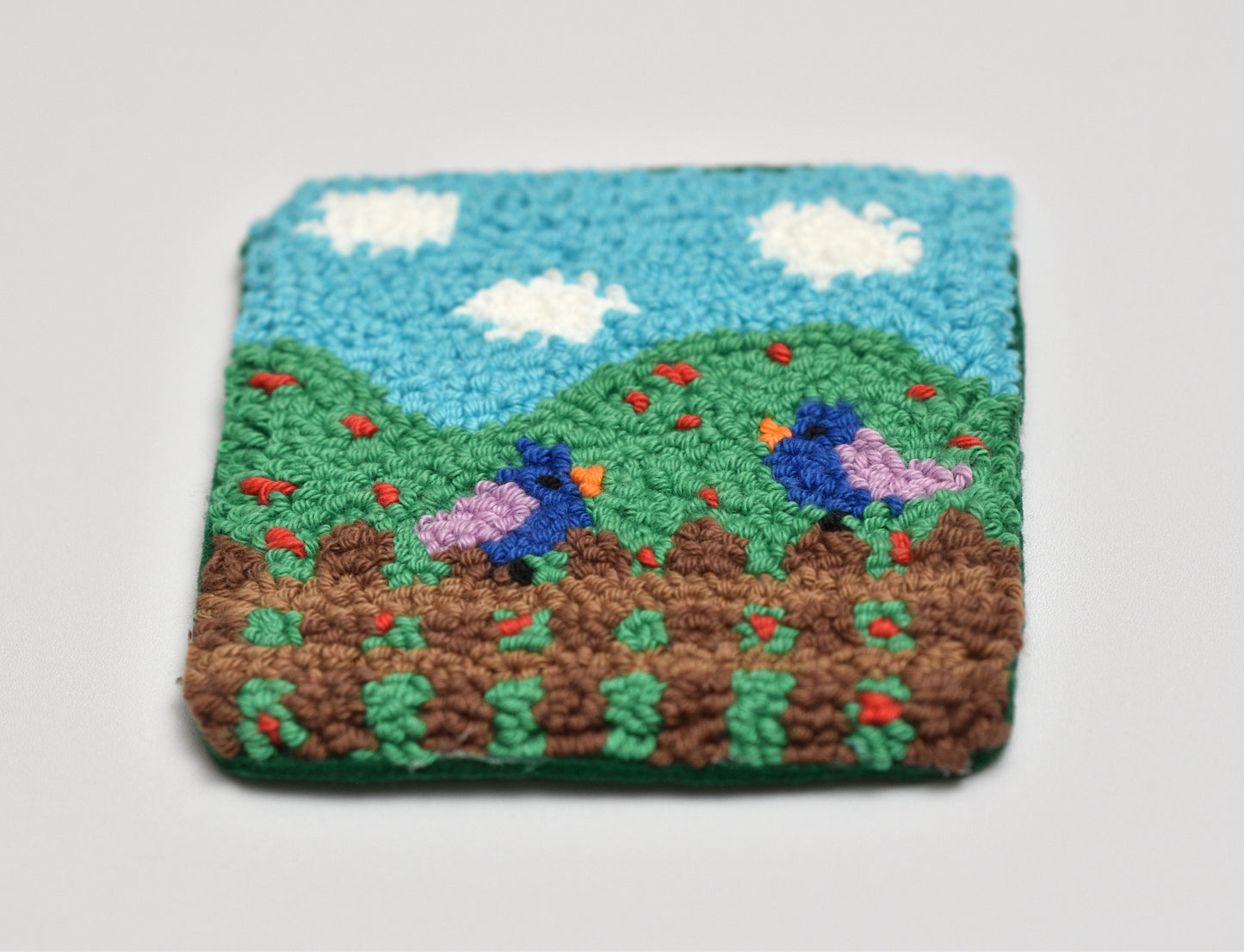 Handmade embroidered landscape with two birds on a green hill under a blue sky with clouds.