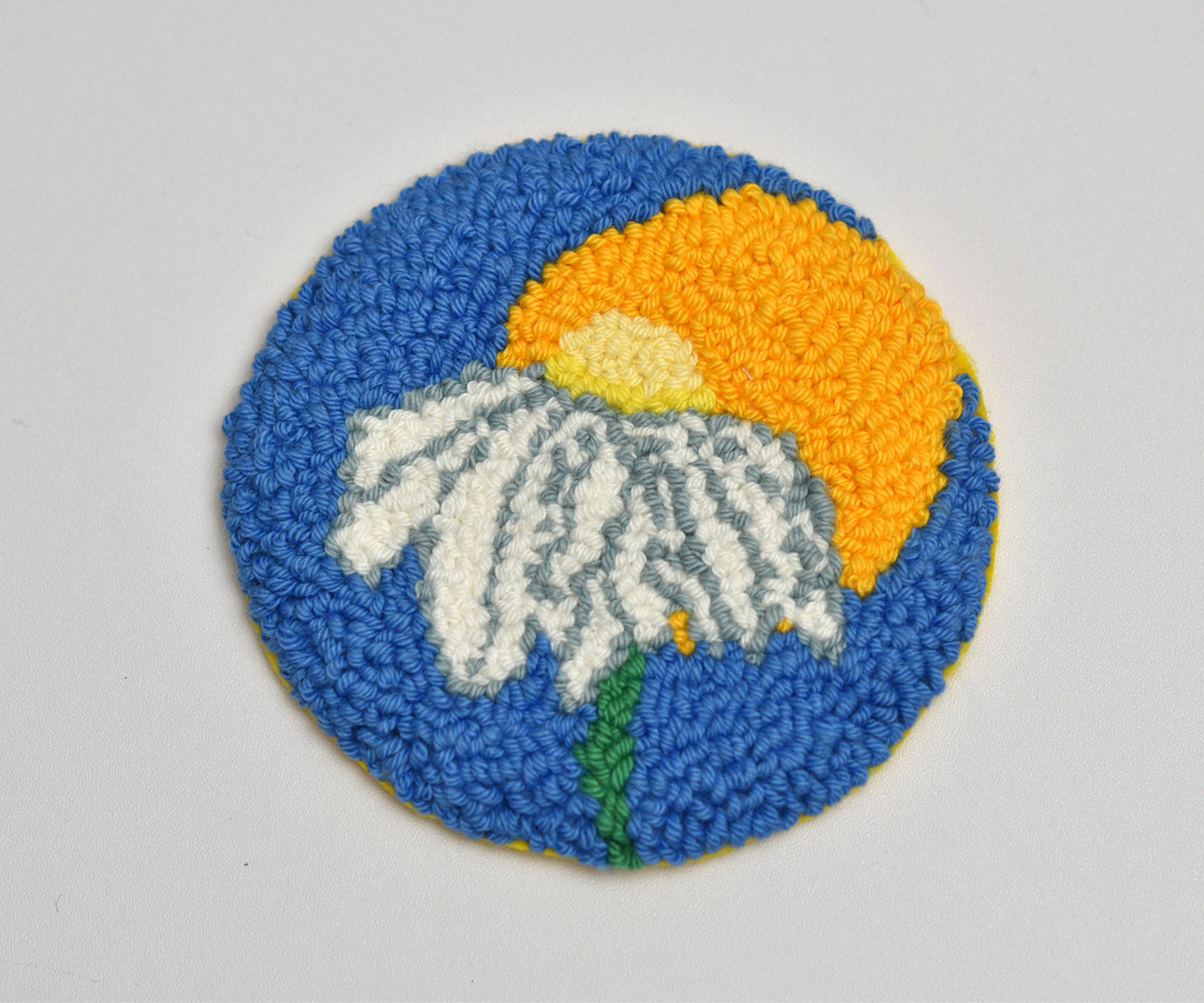 Circular woven coaster with a white daisy flower, blue background, and yellow circle on a gray background