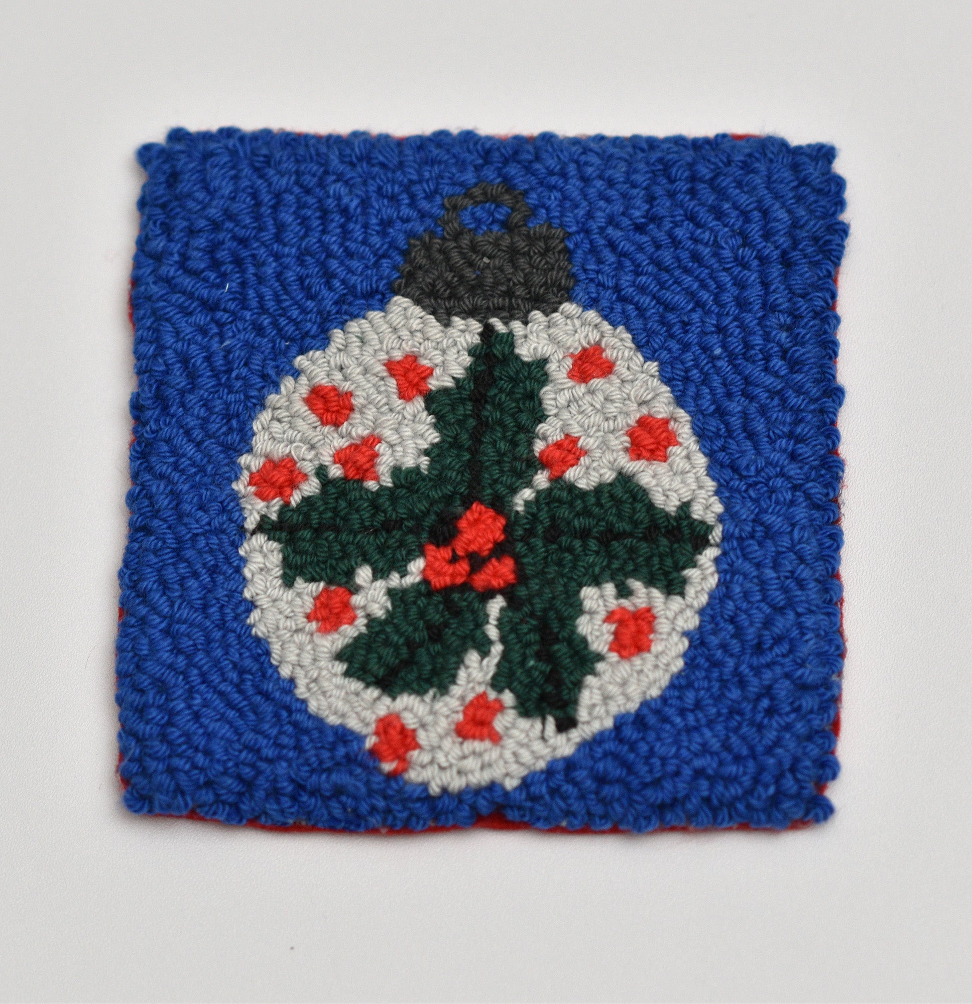 Punch needled coaster with a central design of a Christmas ornament featuring mistle toe holly leaves and berries on a blue background.