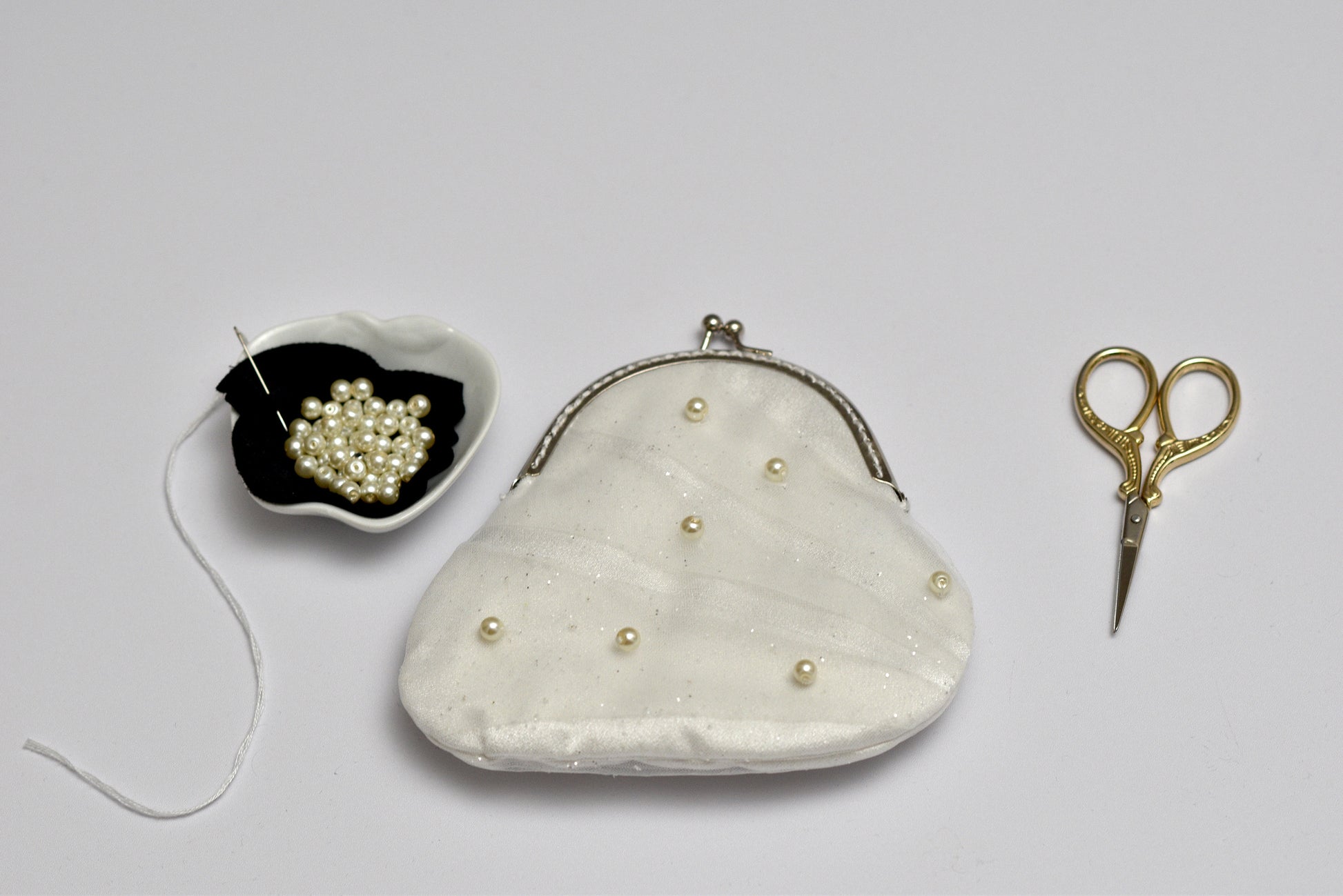 White handbag with pearl embellishments, a small bowl of pearls, and gold scissors on a light gray background