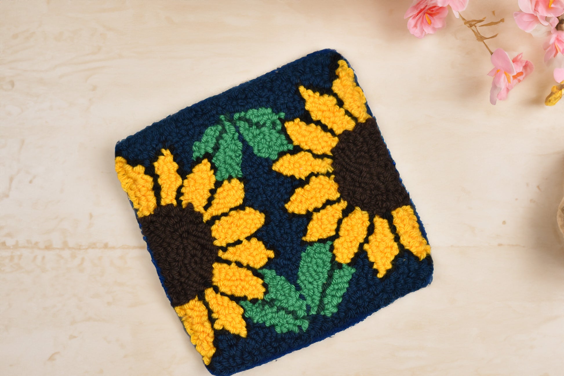 Crocheted sunflower design on a dark blue background with green leaves, placed on a light wooden surface.