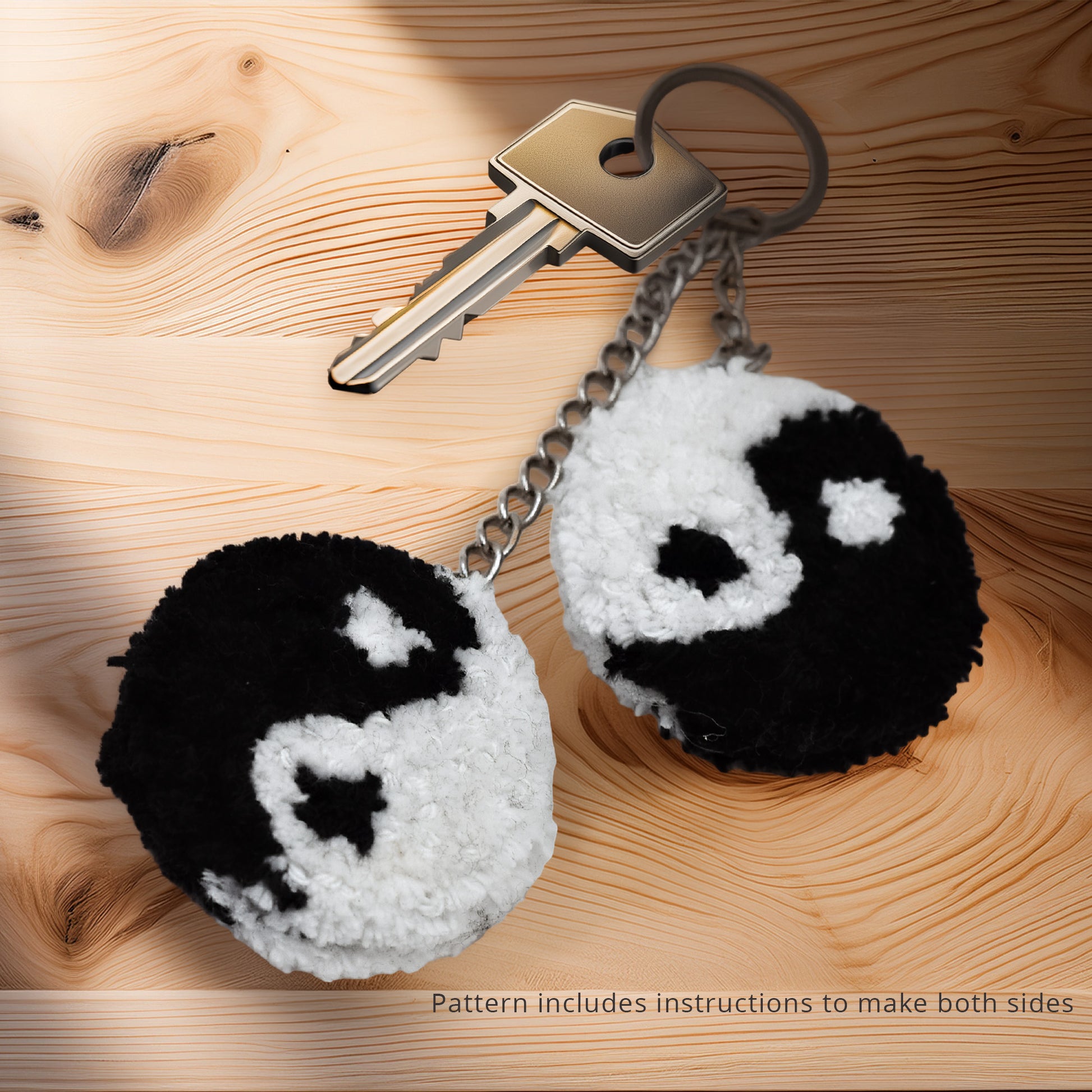 Two black and white yin yang pom pom keychains attached to a silver key on a light wooden surface, with text indicating the pattern includes instructions to make both mirrored sides.
