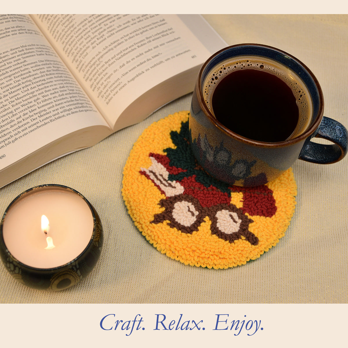 A yellow punch needle coaster with a red book, green maple leaf, and brown framed eye glasses design, shown with a dark ceramic mug filled with coffee, an open book, and a lit candle on a beige fabric surface. The text 'Craft.