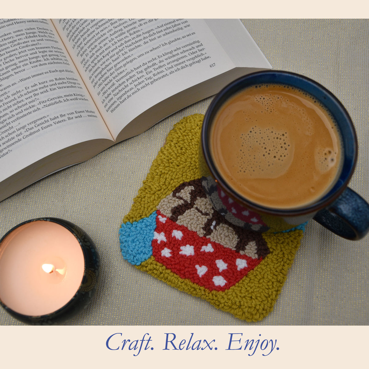 Cherry Top handmade punch needle cupcake coaster beneath a blue coffee mug, styled beside an open book and lit candle, showing the colourful coaster in everyday use.