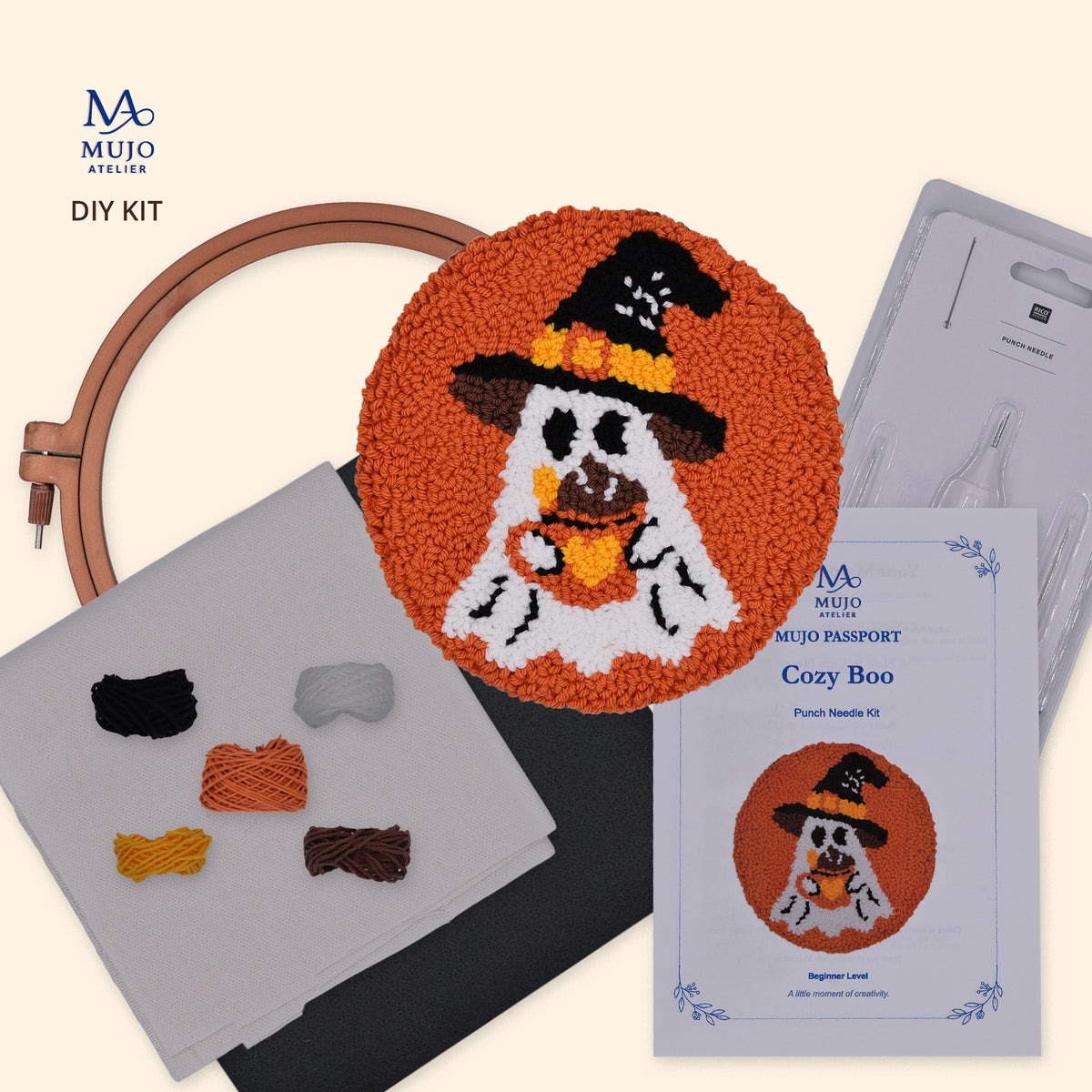 Mujo Atelier DIY Kit for Cozy Boo punch needle kit, featuring a completed orange punch needle coaster with a ghost in a witch hat, a wooden embroidery hoop, yarn samples, a pattern card, and a punch needle on a light beige background.