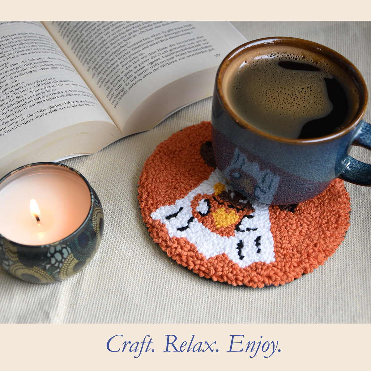 A round orange and white punch needle coaster with a ghost design, shown under a blue ceramic mug filled with coffee. The scene is styled with an open book and a lit candle on a neutral fabric surface, with the text 'Craft.