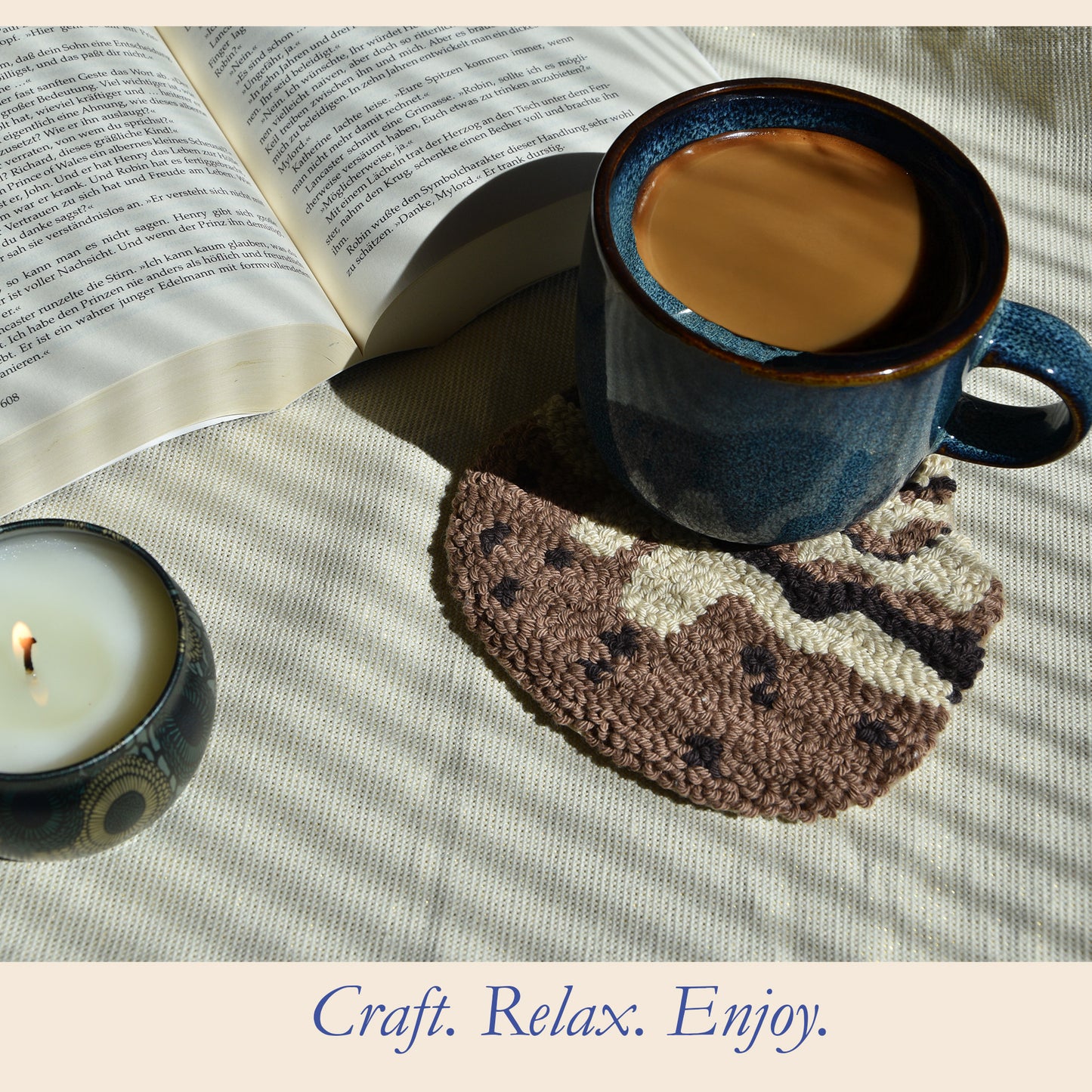 Cozy cinnamon roll punch needle coaster styled with a blue coffee mug, open book and lit candle in warm sunlight, showing the finished Cozy Swirl DIY craft project.