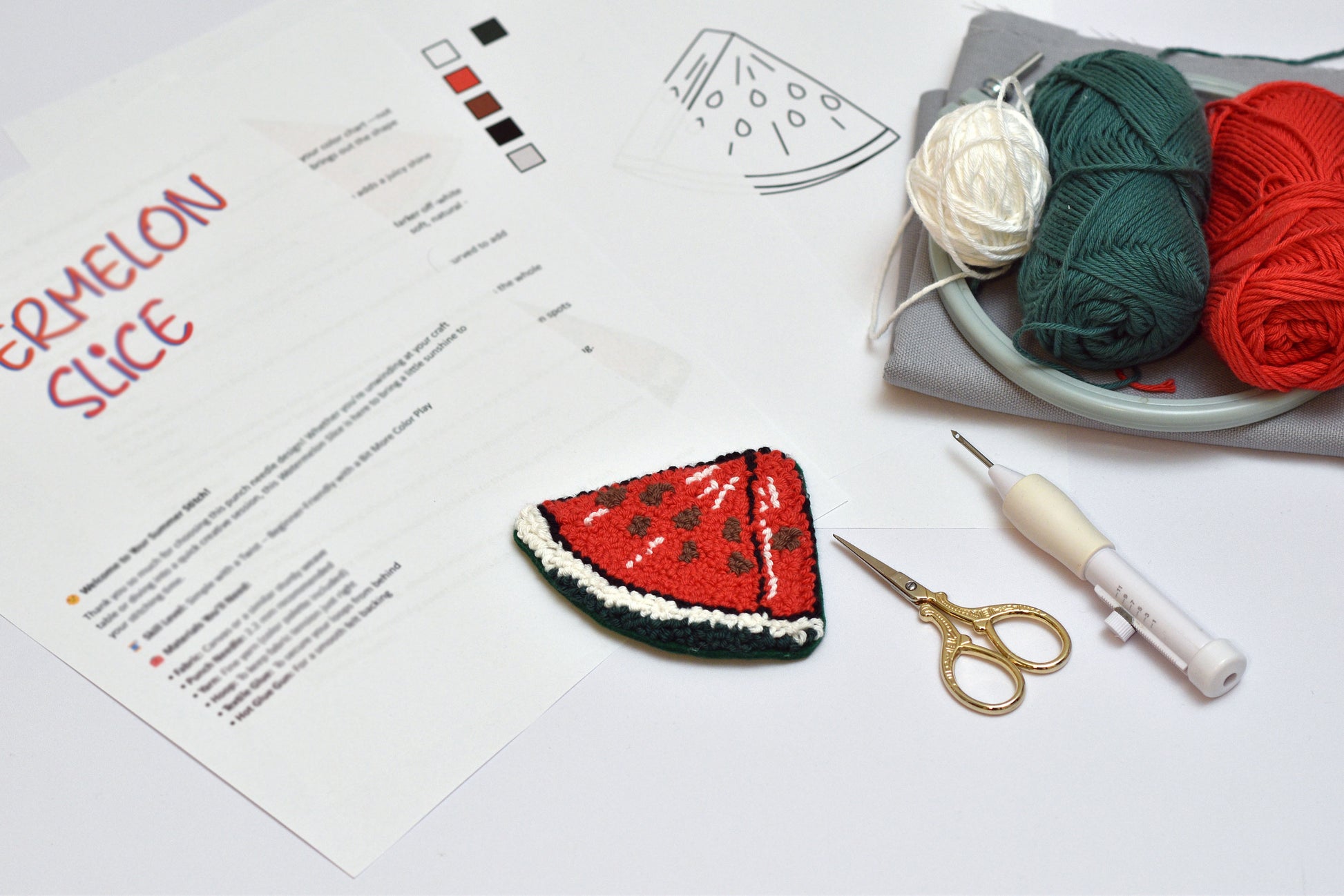Embroidery of a watermelon slice on a white surface with yarn, scissors, and a needle.