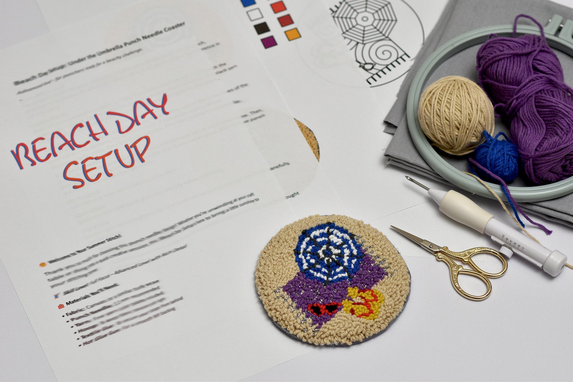 Crafting setup with a completed cross-stitch coaster, yarn, scissors, and a pattern sheet on a white surface.