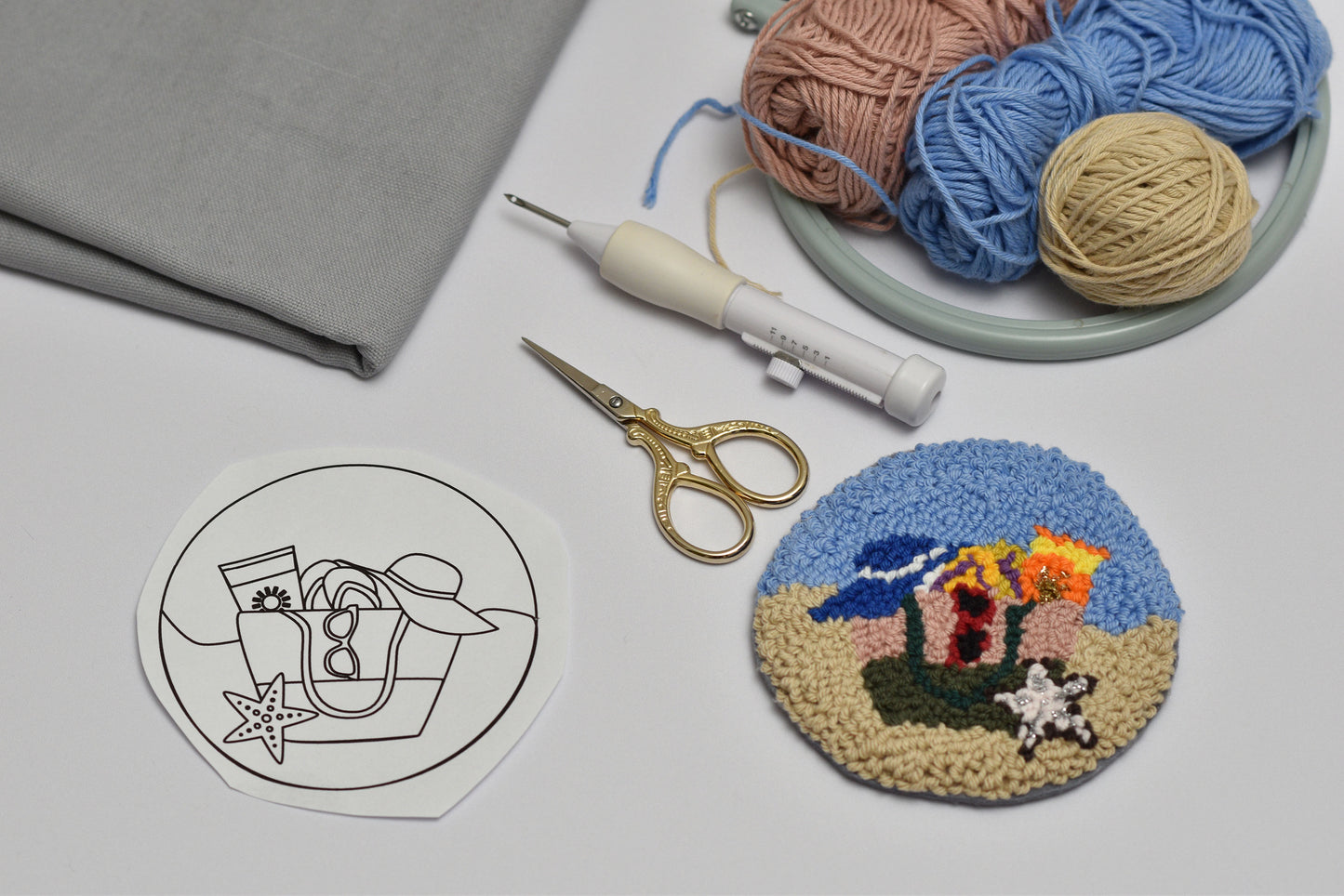 Embroidery hoop with colorful fabric design, yarn balls, scissors, and a printed template on a gray surface.