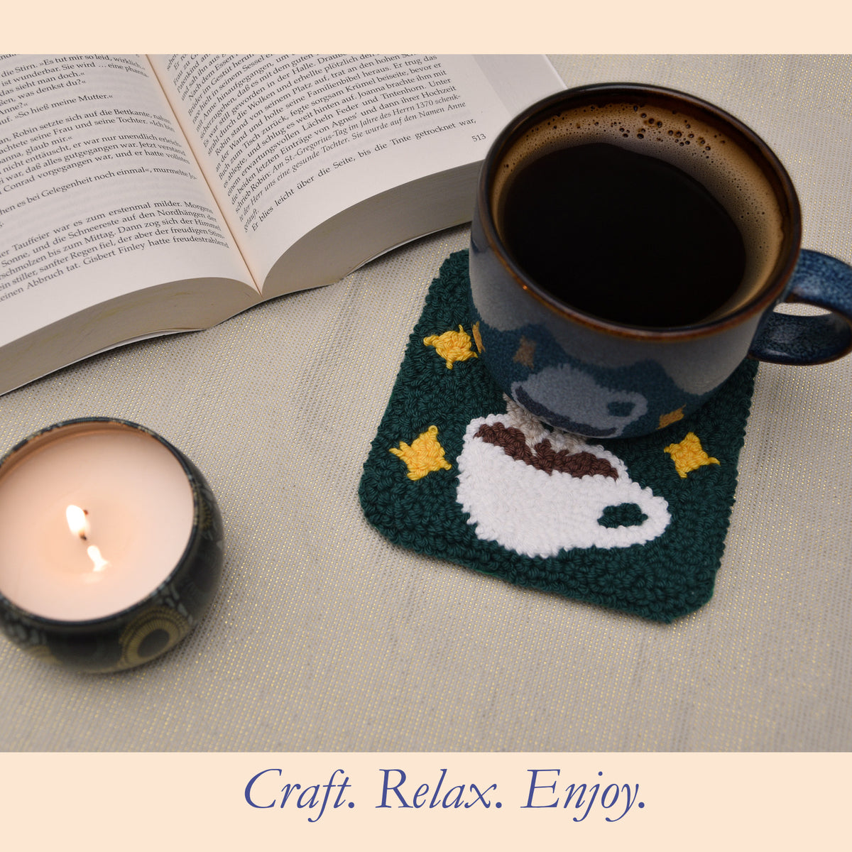 First Light punch needle coffee cup coaster with a dark green background and yellow stars, styled with a blue coffee mug, open book and lit candle in a warm setting.