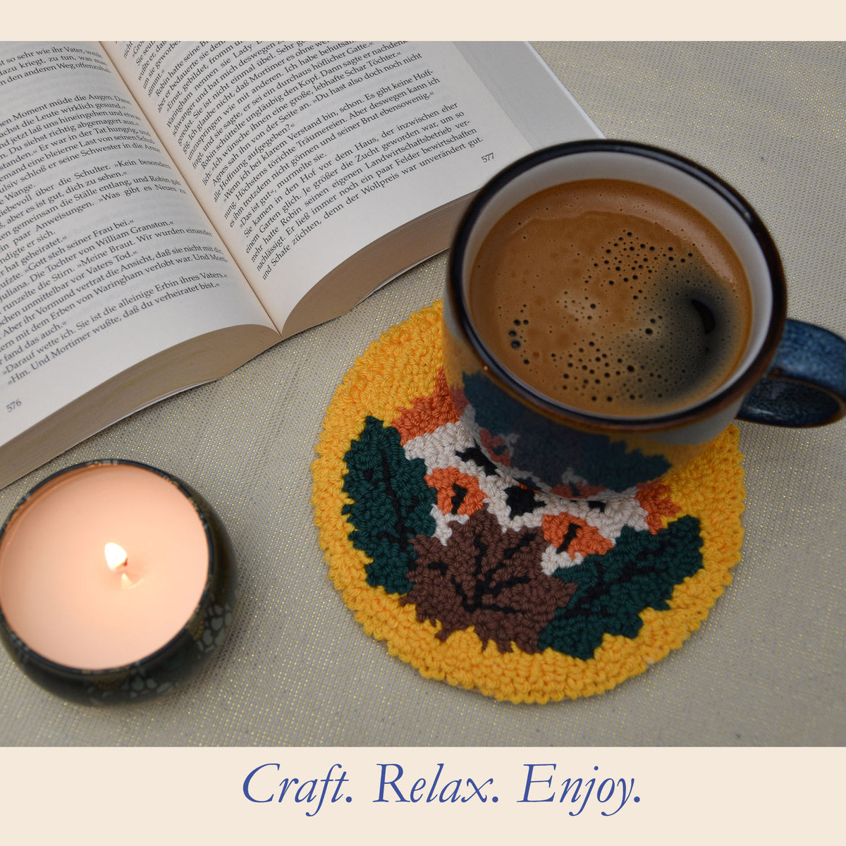 A punch needle circular coaster with a colorful autumn leaf pattern and a fox in yellow, green, brown, and orange, holding a dark blue ceramic mug filled with coffee. An open book and a lit candle are arranged around it on a neutral fabric surface, with the text 'Craft.