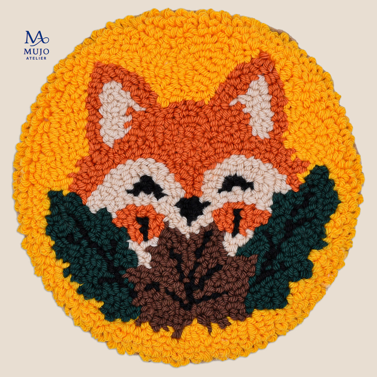 A round, high-pile punch needle coaster featuring a stylized red fox face with dark green and brown leaves at the bottom, set against a bright yellow background. The MUJO ATELIER brand logo is visible in the top left corner.