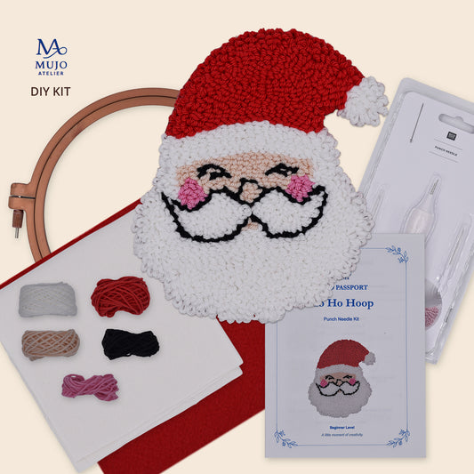 Mujo Atelier DIY Christmas Hoop Art Kit showing a completed Santa face punch needle, embroidery hoop, yarn balls, instruction booklet, and punch needle tools on a light beige background.
