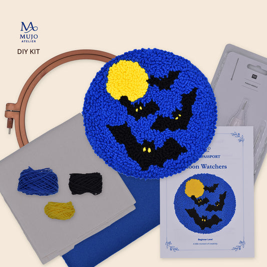 Mujo Atelier DIY punch needle kit featuring a completed blue moon with black bats and a yellow moonstone design, shown alongside embroidery hoops, thread, fabric, a printed pattern sheet, and packaging on a light beige background.