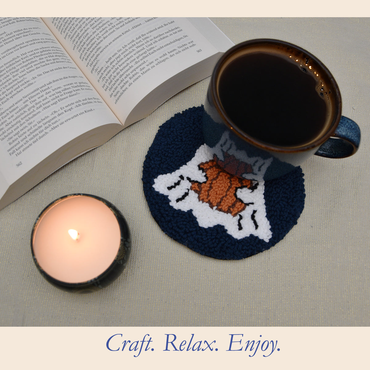 A punch needled mug coaster with a white ghost holding a pumpkin design on a navy blue background shown under a ceramic coffee mug, next to an open book and a lit candle on a neutral fabric surface.