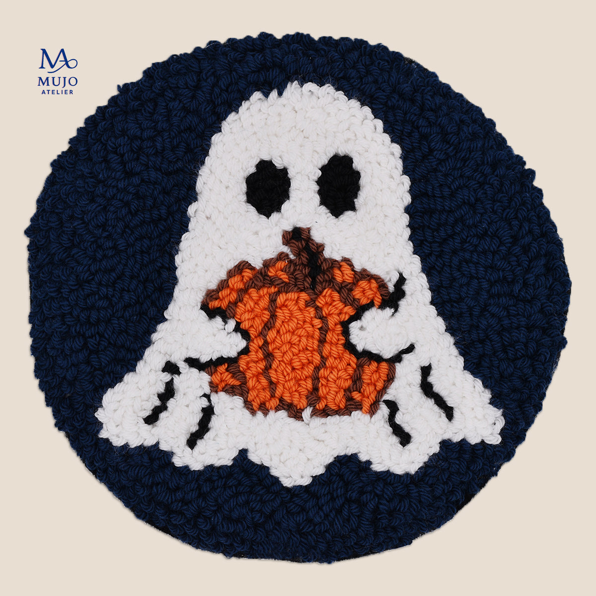 A circular, punch needled Halloween-themed coaster featuring a white ghost holding an orange pumpkin, set against a dark navy blue circular background, shown on a light beige background with a brand watermark in the top left corner.