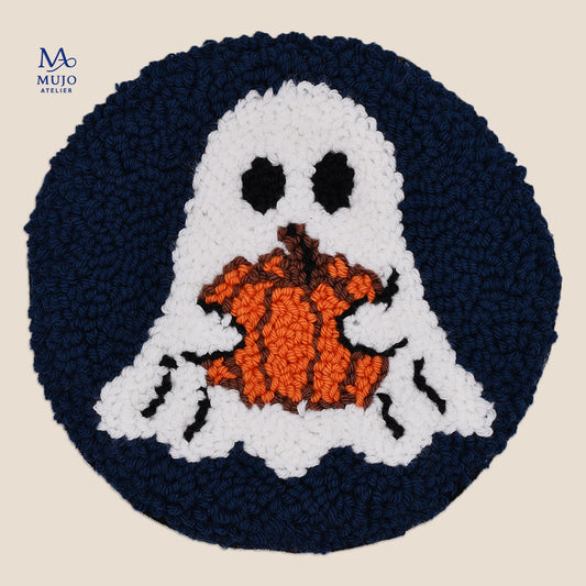 A circular, punch needled Halloween-themed coaster featuring a white ghost holding an orange pumpkin, set against a dark navy blue circular background, shown on a light beige background with a brand watermark in the top left corner.