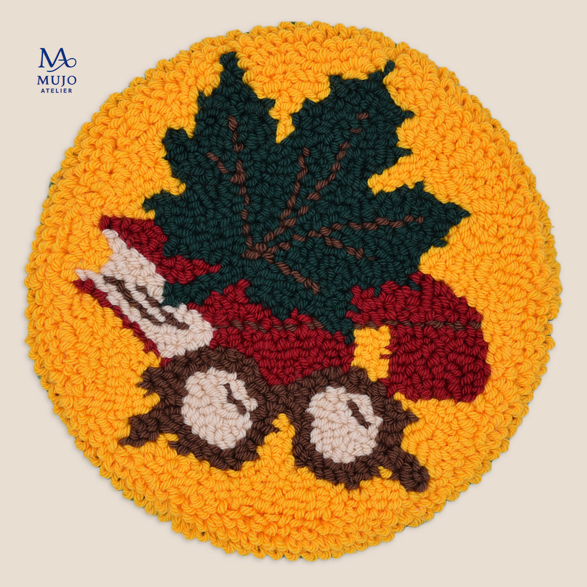 A circular punch needle coaster with a large dark green maple leaf, with brown framed glasses and red book, on a mustard yellow background. The Mujo Atelier logo appears in the upper left corner.